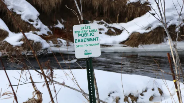 A sign reads “State Fishery Area Public Easement Open to Fishing.” In the background is a stream surrounded by snow on the ground in a natural setting.